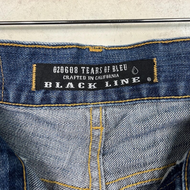 K&ouml;nnte beinhalten: Nahaufnahme einer blauen Jeans mit einem schwarzen Etikett. Auf dem Etikett steht "020608 TEARS OF BLEU CRAFTED IN CALIFORNIA BLACK LINE". Die Jeans hat gelbe N&auml;hte und ein strukturiertes Aussehen.
