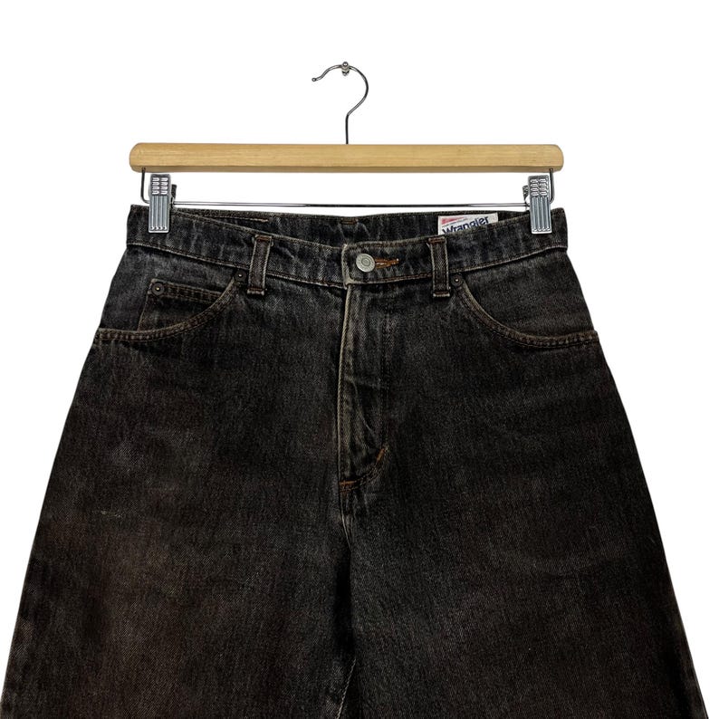 Puede incluir: Vaqueros Wrangler de mezclilla negra colgados de una percha de madera. Los vaqueros tienen un dise&ntilde;o cl&aacute;sico de cinco bolsillos, cierre de bot&oacute;n y cremallera, y el logotipo de Wrangler. Los vaqueros son de lavado oscuro con sutiles desvanecimientos.