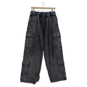 Könnte beinhalten: Grau gewaschene Cargo-Hose mit mehreren Taschen und einem Kordelzug in der Taille. Die Hose mit weitem Bein hängt an einem Holzbügel vor weißem Hintergrund. Die Hose hat ein verwaschenes, strukturiertes Aussehen.