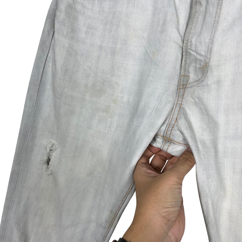 May include: Light wash denim jeans with a distressed knee and visible wear. The jeans are held by a hand, showcasing the light blue color and the texture of the fabric. The stitching is a contrasting tan color.