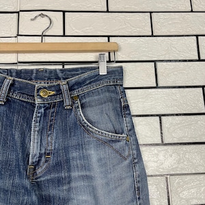 May include: Close-up of blue denim jeans hanging on a wooden hanger. The jeans have a button closure, front pockets, and visible stitching. The denim has a faded appearance, with lighter areas on the front.