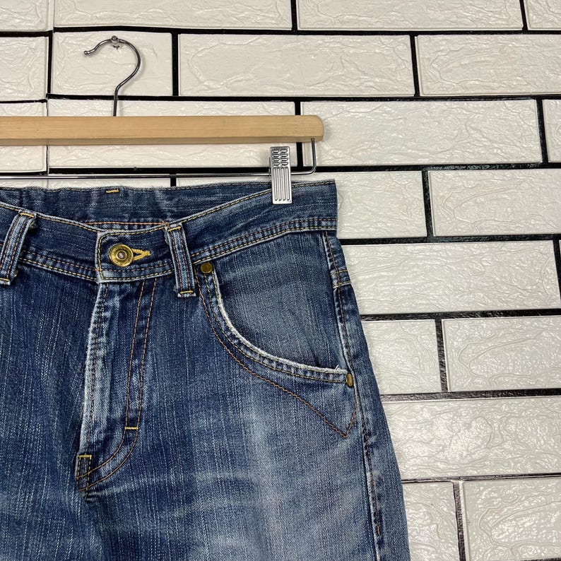 May include: Close-up of blue denim jeans hanging on a wooden hanger. The jeans have a button closure, front pockets, and visible stitching. The denim has a faded appearance, with lighter areas on the front.