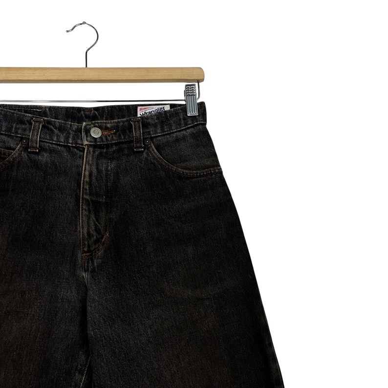 Puede incluir: Vaqueros de mezclilla de lavado oscuro colgados de una percha de madera. Los vaqueros tienen un dise&ntilde;o cl&aacute;sico de cinco bolsillos y cierre de bot&oacute;n y cremallera. La marca "Wrangler" es visible en la cintura.