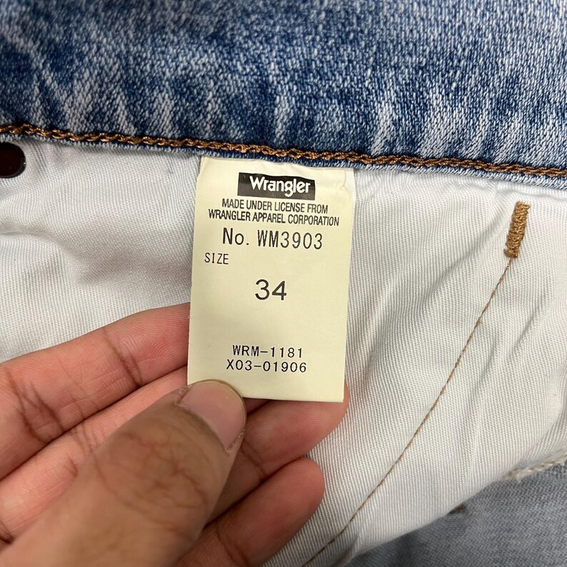 K&ouml;nnte beinhalten: Nahaufnahme eines Wrangler Jeans-Etiketts. Das Etikett ist wei&szlig; mit schwarzem Text, einschlie&szlig;lich des Markennamens, der Gr&ouml;&szlig;e 34 und Stilinformationen. Die Jeans ist aus hellblauem Denim.