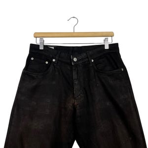 May include: Black denim jeans hanging on a wooden hanger. The jeans have a classic five-pocket design with a button and zipper closure. The denim has a faded, worn appearance.