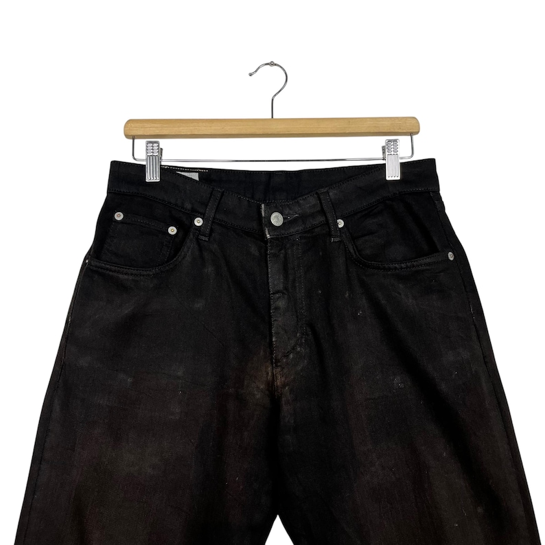 May include: Black denim jeans hanging on a wooden hanger. The jeans have a classic five-pocket design with a button and zipper closure. The denim has a faded, worn appearance.