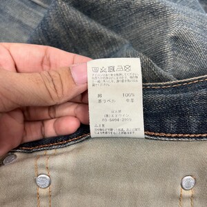 May include: Close-up of a denim garment with a white tag. The tag has Japanese text and care symbols. The denim is a faded blue color. The tag also has the words "100%" and "leather label".