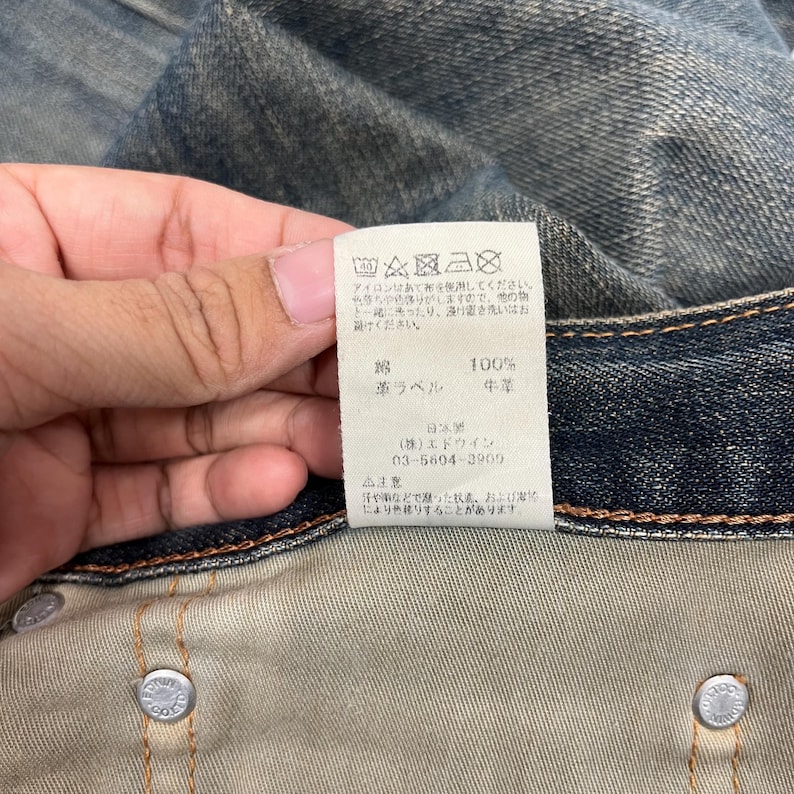 May include: Close-up of a denim garment with a white tag. The tag has Japanese text and care symbols. The denim is a faded blue color. The tag also has the words "100%" and "leather label".