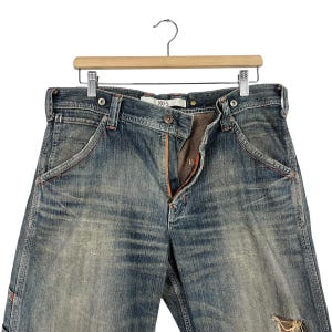 May include: Blue denim jeans with distressed details, hanging on a wooden hanger. The jeans feature orange stitching, a button closure, and a zipper. The brand name "XUS" is visible on the waistband.