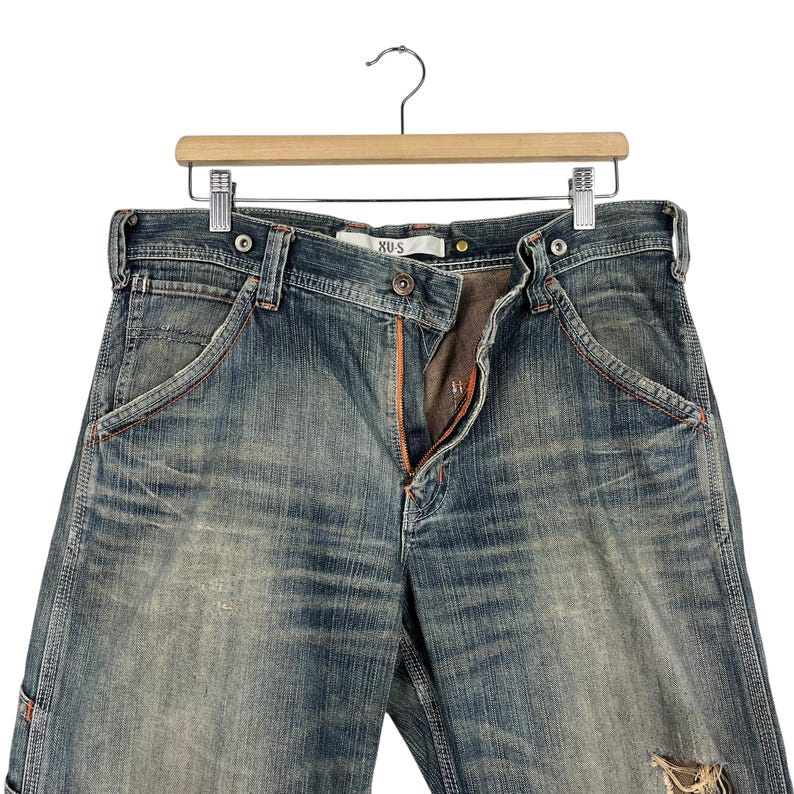 May include: Blue denim jeans with distressed details, hanging on a wooden hanger. The jeans feature orange stitching, a button closure, and a zipper. The brand name "XUS" is visible on the waistband.