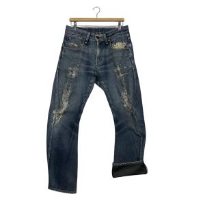 K&ouml;nnte beinhalten: Blaue Jeans aus Denim mit Used-Details, h&auml;ngen an einem Holzb&uuml;gel. Die Jeans haben eine verblasste Waschung mit wei&szlig;en Akzenten und einen umgeschlagenen Saum. Die Jeans sind im klassischen F&uuml;nf-Taschen-Stil.