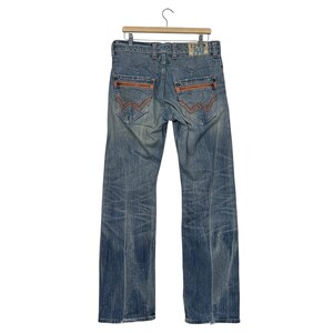 May include: Faded blue denim jeans with orange stitching details on the back pockets and seams. The jeans are hanging on a wooden hanger. The jeans have a straight leg cut.