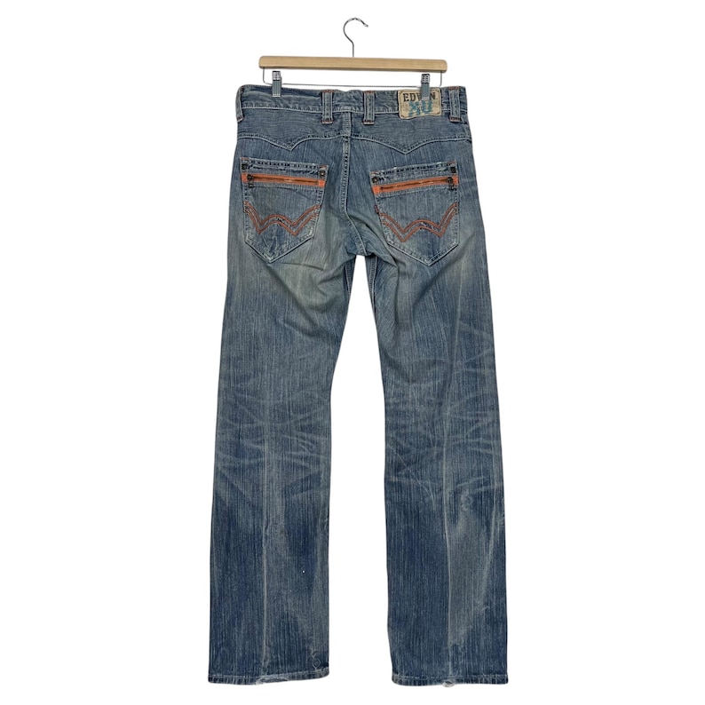 May include: Faded blue denim jeans with orange stitching details on the back pockets and seams. The jeans are hanging on a wooden hanger. The jeans have a straight leg cut.