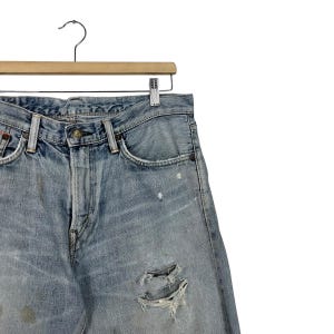 May include: Light blue denim jeans with distressed details, including rips and faded patches. The jeans are hanging on a wooden hanger with a metal clip. The jeans have a classic five-pocket design.