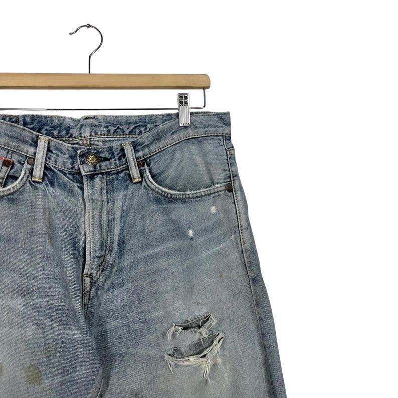 May include: Light blue denim jeans with distressed details, including rips and faded patches. The jeans are hanging on a wooden hanger with a metal clip. The jeans have a classic five-pocket design.