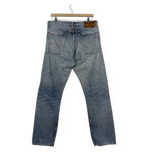 May include: Light blue denim jeans with a faded wash and distressed details. The jeans are hanging on a wooden hanger. The back pockets and leather patch are visible. The jeans have a straight leg cut.