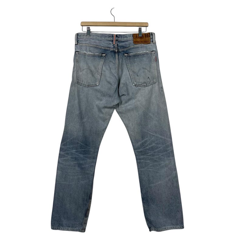 May include: Light blue denim jeans with a faded wash and distressed details. The jeans are hanging on a wooden hanger. The back pockets and leather patch are visible. The jeans have a straight leg cut.