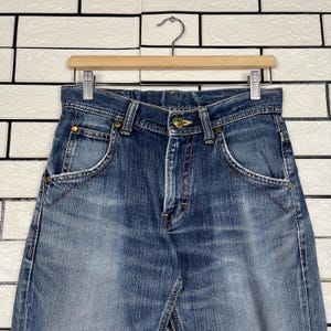 May include: Blue denim jeans with a classic five-pocket design, featuring a button closure and brass rivets. The jeans are hanging on a wooden hanger against a white brick wall background. The denim has a faded wash.