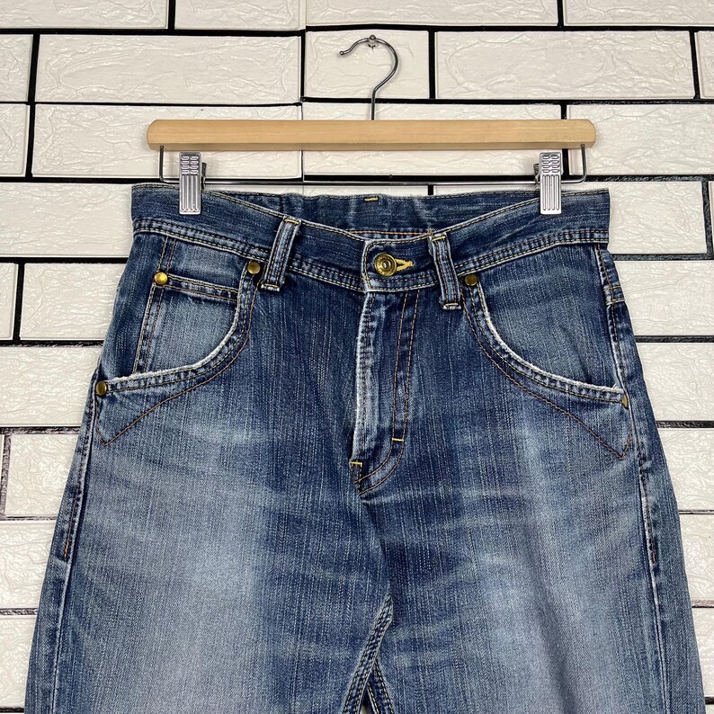 May include: Blue denim jeans with a classic five-pocket design, featuring a button closure and brass rivets. The jeans are hanging on a wooden hanger against a white brick wall background. The denim has a faded wash.