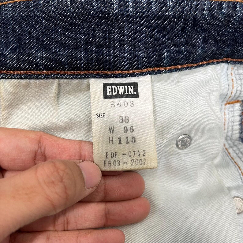K&ouml;nnte beinhalten: Nahaufnahme eines Jeansetiketts mit der Marke "Edwin". Das Etikett zeigt die Gr&ouml;&szlig;e 38, mit den Ma&szlig;en W 96 cm und H 113 cm. Das Etikett enth&auml;lt auch die Codes S403, EDF-0712 und E503-2002.