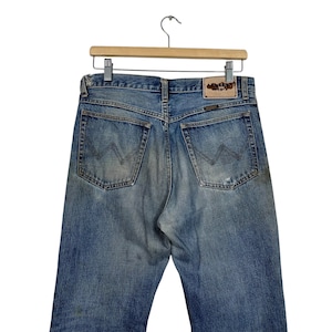 K&ouml;nnte beinhalten: Hellblaue Jeans mit klassischem F&uuml;nf-Taschen-Design und dem Wrangler-Logo auf einem Lederpatch. Die Jeans sind abgenutzt mit ausgeblichenen Bereichen, h&auml;ngen an einem Holzkleiderb&uuml;gel.