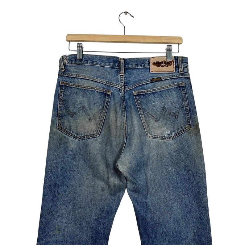 K&ouml;nnte beinhalten: Hellblaue Jeans mit klassischem F&uuml;nf-Taschen-Design und dem Wrangler-Logo auf einem Lederpatch. Die Jeans sind abgenutzt mit ausgeblichenen Bereichen, h&auml;ngen an einem Holzkleiderb&uuml;gel.