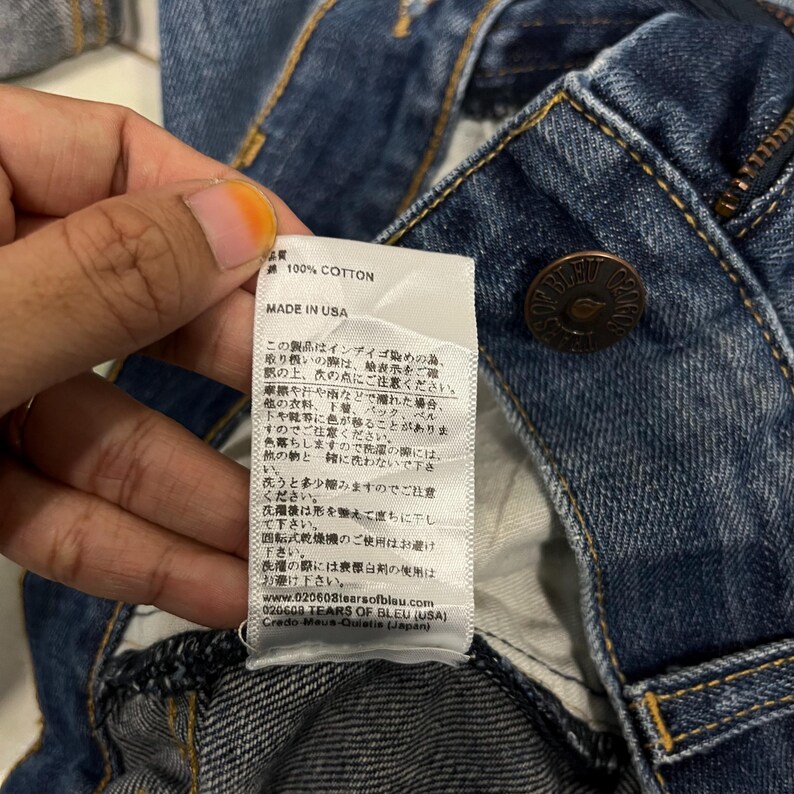 K&ouml;nnte beinhalten: Nahaufnahme einer blauen Jeans mit Knopf- und Rei&szlig;verschluss. Ein wei&szlig;es Etikett mit japanischem Text und den Worten "100% COTTON" und "MADE IN USA" ist sichtbar. Die Jeans besteht aus strapazierf&auml;higem Baumwollmaterial.