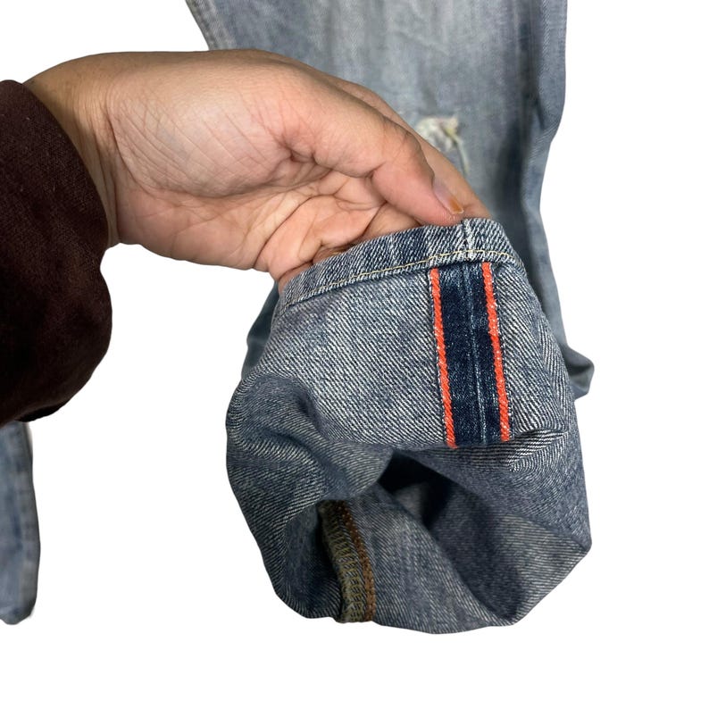 May include: Close-up of light blue denim jeans. The cuff is turned up, revealing a dark blue and orange stripe detail. The jeans have a distressed look with some visible wear and tear.