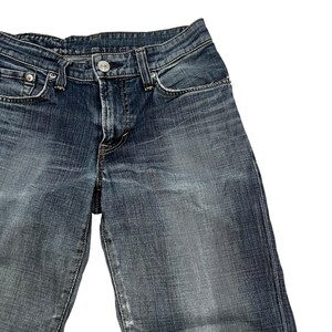 Op de afbeelding: Close-up van een blauwe spijkerbroek. De jeans heeft een klassiek ontwerp met vijf zakken, een knoop- en ritssluiting en een vervaagde wassing. De denimstof vertoont tekenen van slijtage, wat bijdraagt aan de vintage esthetiek.
