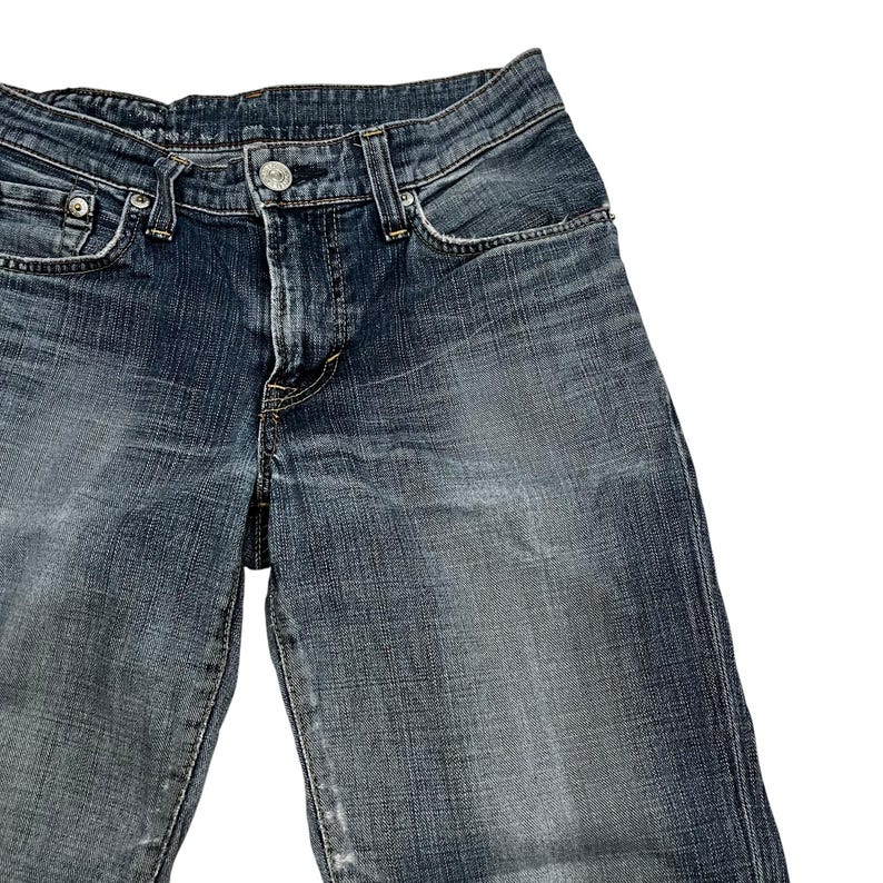 Op de afbeelding: Close-up van een blauwe spijkerbroek. De jeans heeft een klassiek ontwerp met vijf zakken, een knoop- en ritssluiting en een vervaagde wassing. De denimstof vertoont tekenen van slijtage, wat bijdraagt aan de vintage esthetiek.