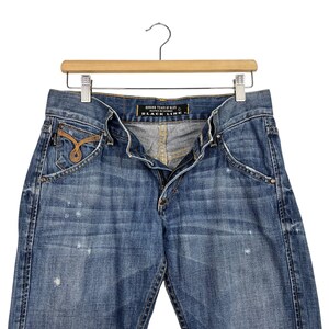 K&ouml;nnte beinhalten: Blaue Jeans aus Denim mit einem Used-Look, h&auml;ngen an einem Holzb&uuml;gel. Die Jeans haben einen Knopf- und Rei&szlig;verschluss, Vordertaschen und ein dekoratives goldfarbenes Design auf der Vordertasche. Der Text "GODDESS TEARS OF BLUE BLACK LINE" ist sichtbar.