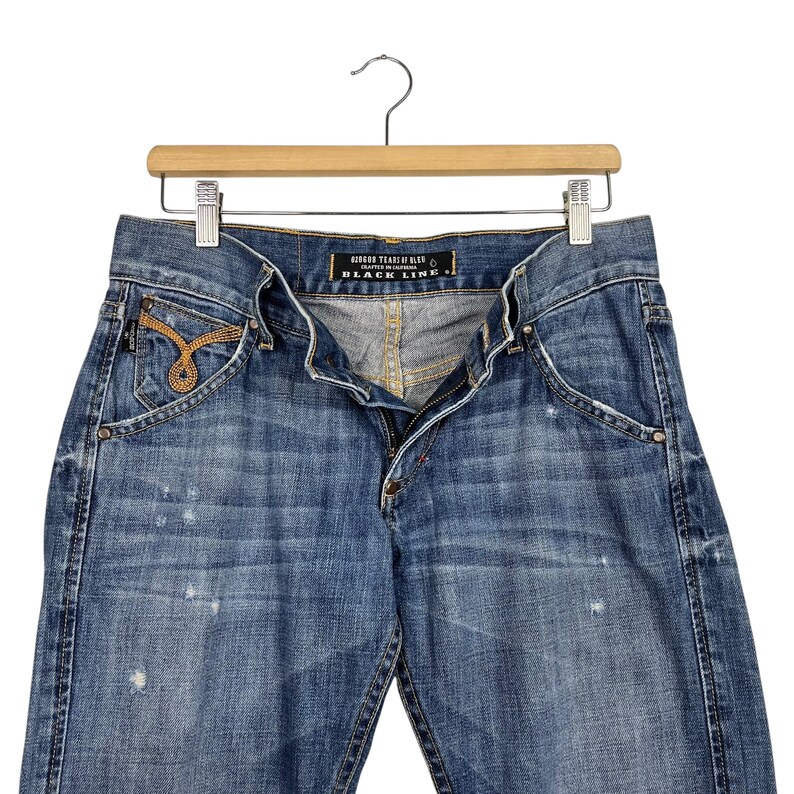K&ouml;nnte beinhalten: Blaue Jeans aus Denim mit einem Used-Look, h&auml;ngen an einem Holzb&uuml;gel. Die Jeans haben einen Knopf- und Rei&szlig;verschluss, Vordertaschen und ein dekoratives goldfarbenes Design auf der Vordertasche. Der Text "GODDESS TEARS OF BLUE BLACK LINE" ist sichtbar.