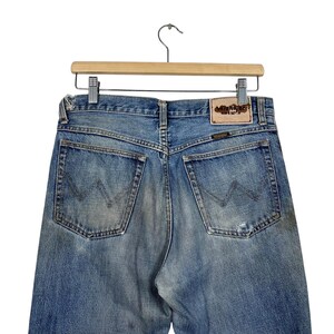 K&ouml;nnte beinhalten: Hellblaue Jeans mit Used-Look, h&auml;ngen an einem Holzb&uuml;gel. Die Ges&auml;&szlig;taschen haben ein aufgesticktes Design. Ein Lederpatch mit Text ist am Bund sichtbar.