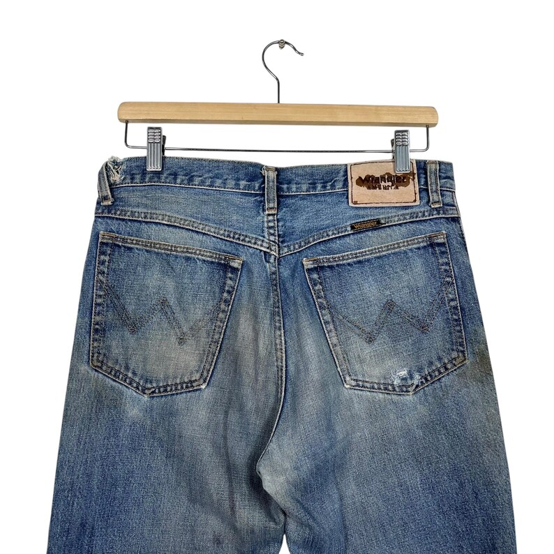 K&ouml;nnte beinhalten: Hellblaue Jeans mit Used-Look, h&auml;ngen an einem Holzb&uuml;gel. Die Ges&auml;&szlig;taschen haben ein aufgesticktes Design. Ein Lederpatch mit Text ist am Bund sichtbar.