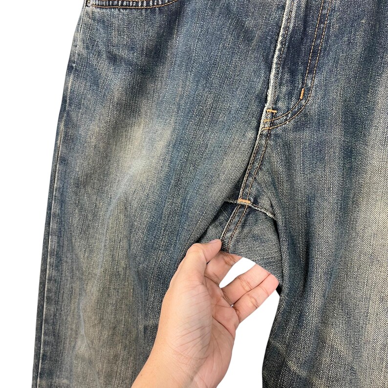 May include: Close-up of blue denim jeans with visible stitching and a faded wash. The jeans are held open, showcasing the fabric's texture and construction details. The denim has a worn appearance.