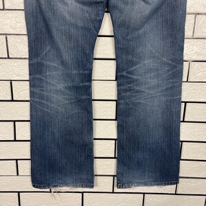 May include: A pair of blue denim jeans with a straight leg cut. The jeans have a faded wash effect and visible wear marks. The background is a white brick wall.