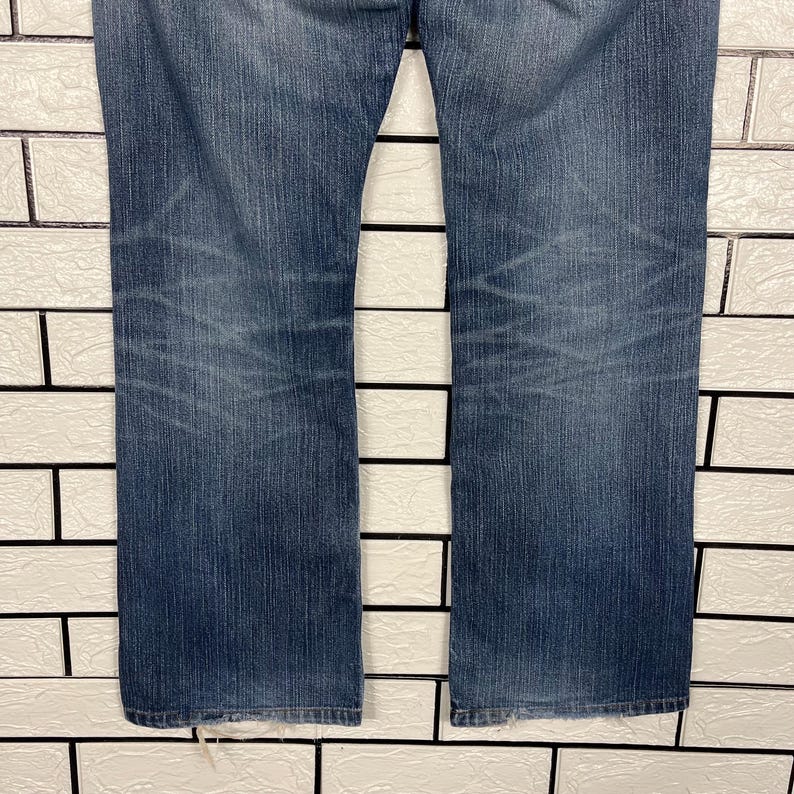 May include: A pair of blue denim jeans with a straight leg cut. The jeans have a faded wash effect and visible wear marks. The background is a white brick wall.