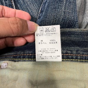 May include: Close-up of a denim jeans tag with Japanese text and care symbols. The tag indicates the jeans are 100% cotton with a synthetic leather label. The tag also includes the text "MADE IN CHINA".