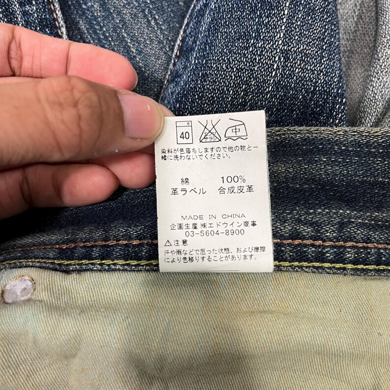 May include: Close-up of a denim jeans tag with Japanese text and care symbols. The tag indicates the jeans are 100% cotton with a synthetic leather label. The tag also includes the text "MADE IN CHINA".