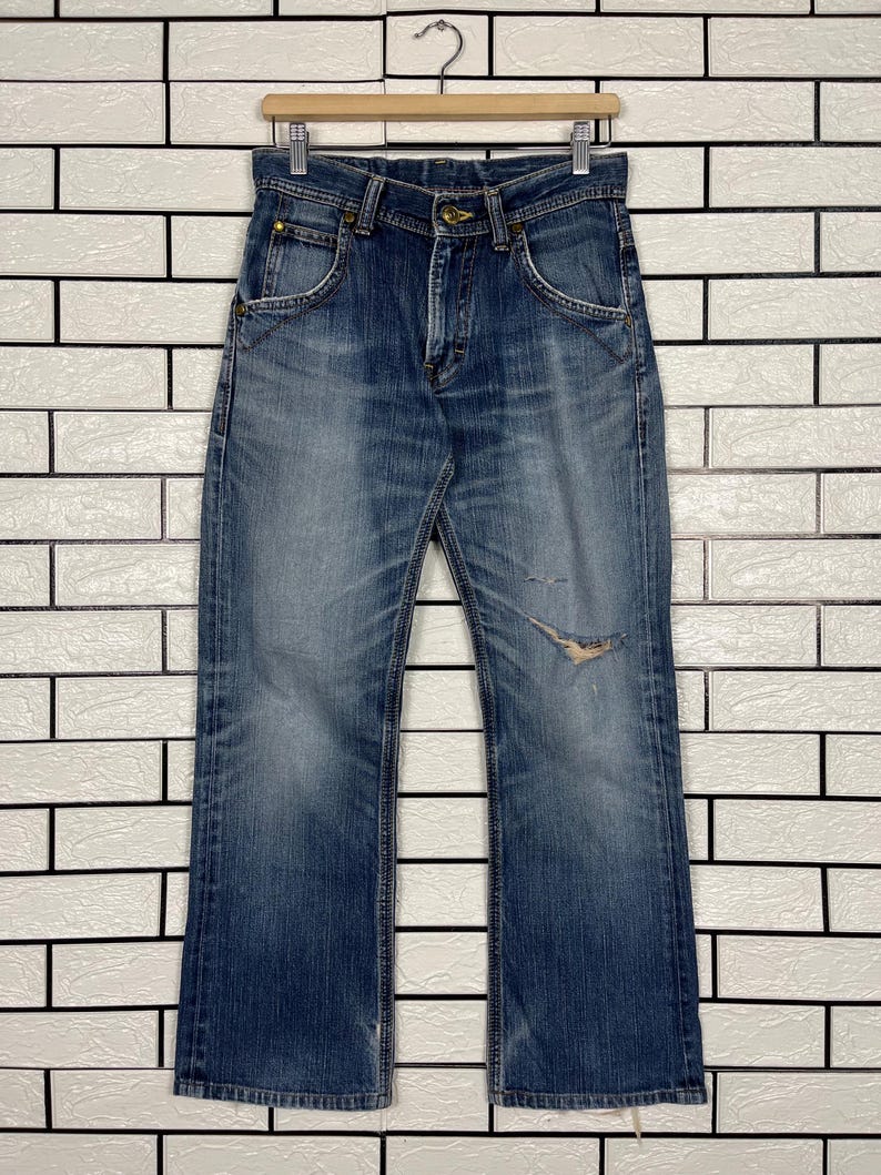 May include: Faded blue denim jeans with a distressed knee detail, hanging on a wooden hanger. The jeans have a classic five-pocket design and a slightly flared leg. The denim shows signs of wear and fading.