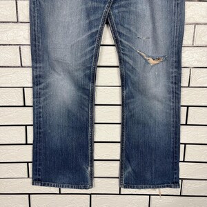May include: Faded blue denim jeans with a distressed, worn look. The jeans have a classic five-pocket design, a button and zipper closure, and a slight bootcut leg. There is a large tear on the right leg, adding to the vintage aesthetic.