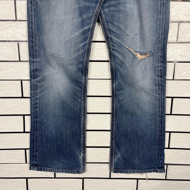 May include: Faded blue denim jeans with a distressed, worn look. The jeans have a classic five-pocket design, a button and zipper closure, and a slight bootcut leg. There is a large tear on the right leg, adding to the vintage aesthetic.