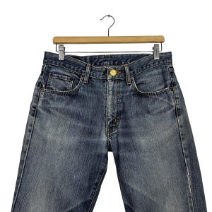 May include: Close-up of blue denim jeans hanging on a wooden hanger. The jeans have a classic five-pocket design, a button closure, and a zipper fly. The denim has a faded, worn look.