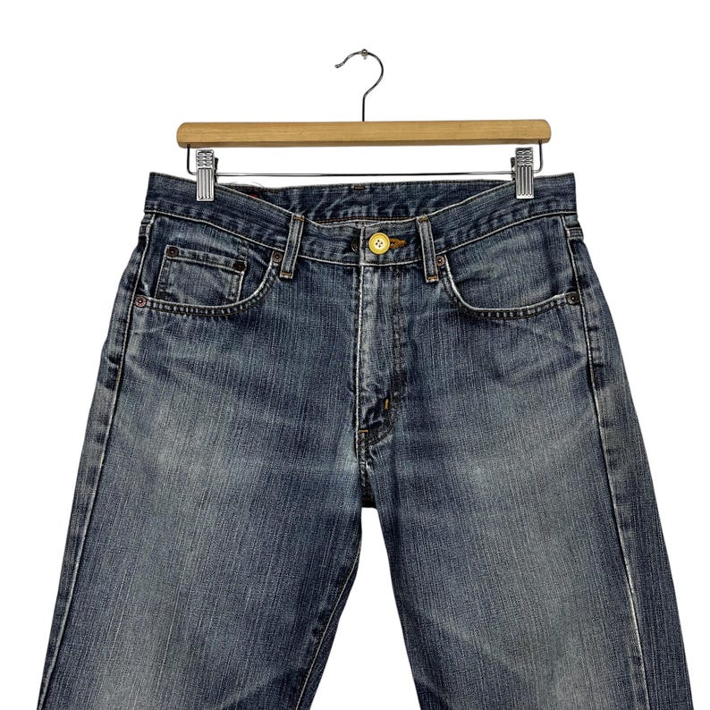 May include: Close-up of blue denim jeans hanging on a wooden hanger. The jeans have a classic five-pocket design, a button closure, and a zipper fly. The denim has a faded, worn look.