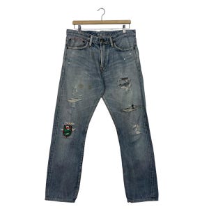 May include: Light blue denim jeans with distressed details, including rips and patches. The jeans are hanging on a wooden hanger. A colorful patch is visible on the left leg.