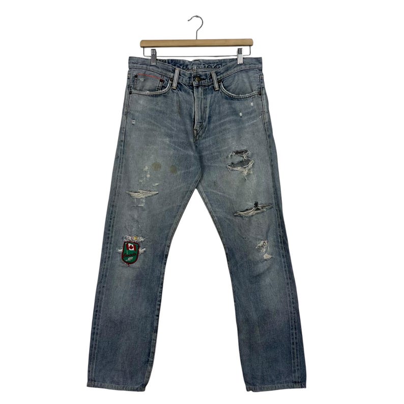 May include: Light blue denim jeans with distressed details, including rips and patches. The jeans are hanging on a wooden hanger. A colorful patch is visible on the left leg.