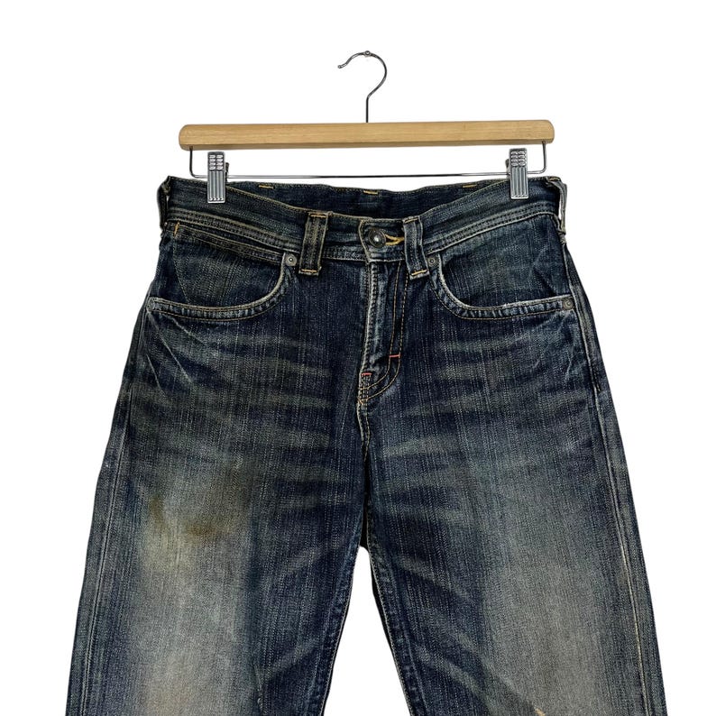 May include: Dark wash denim jeans hanging on a wooden hanger. The jeans have a classic five-pocket design, a button and zipper closure, and show signs of wear with faded areas and subtle distressing. The jeans are a medium rise and have a straight leg.