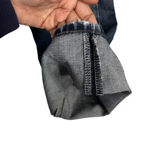 May include: Close-up of the hem of blue denim jeans, folded up to show the inside fabric and stitching. The jeans appear worn, with some fraying at the hem. The denim is a classic blue colour.