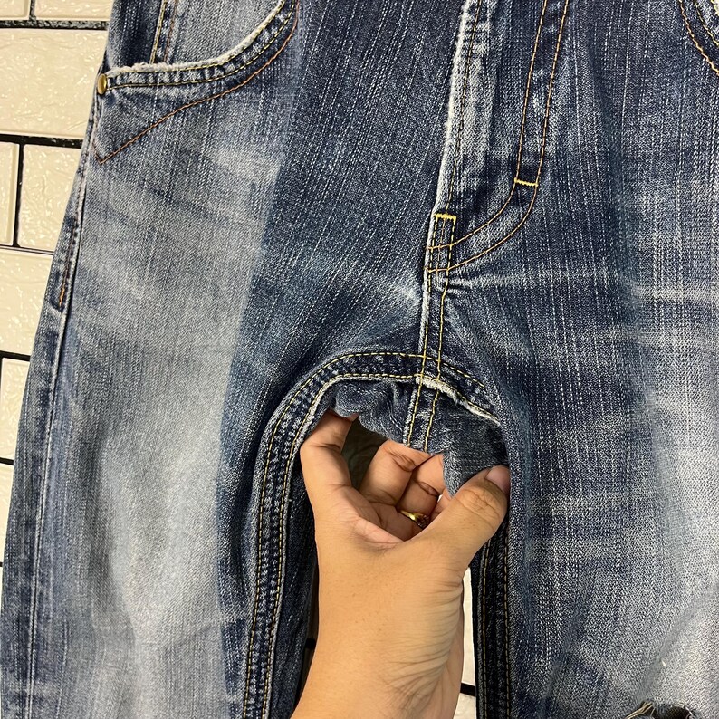 May include: Close-up of blue denim jeans with visible stitching and pocket details. The jeans show signs of wear and fading, with a hand holding the fabric. The denim has a classic, casual style.