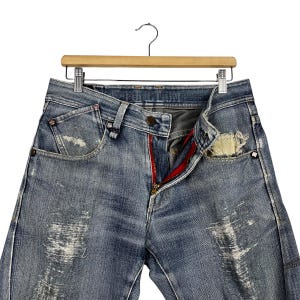 K&ouml;nnte beinhalten: Hellblaue Jeans mit Used-Details, rotem Rei&szlig;verschluss und Knopfverschluss. Die Jeans h&auml;ngen an einem Holzb&uuml;gel. Die Taschen haben einen abgenutzten Look.